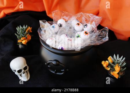 Calderone nero Witcher pieno di palline oculari di plastica come ingredienti per la pozione magica. Decorazione di Halloween con teschio e fiori di cactus. Foto Stock