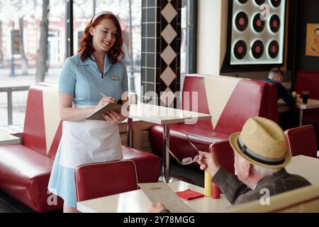 Ritratto di cameriera sorridente in uniforme blu brillante che prende un ordine da un anziano gentiluomo in un ristorante retrò con cabine rosse e decorazioni con dischi in vinile Foto Stock