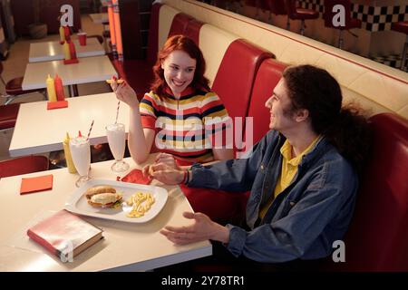 Una giovane coppia sorride e si gusta il pasto in uno stand retrò, con frullati di latte, patatine fritte e un hamburger sul tavolo, facendo una piacevole conversazione Foto Stock