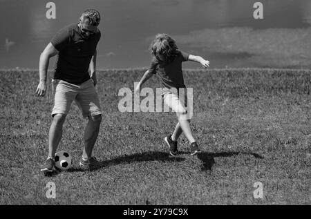 Padre con figlio che gioca a calcio sul campo. Padre e figlio giocano a calcio insieme nel parco estivo. Il figlio eccitatamente calcia la palla verso il padre Foto Stock