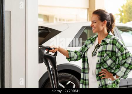 Donna in una stazione di ricarica per veicoli elettrici in città che carica la sua auto elettrica. Energia pulita, elettrificazione e consapevolezza ambientale. Foto Stock