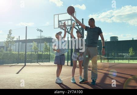 una famiglia sportiva gioca a basket, padre, figlie trascorrono il tempo libero sul campo da basket, conducono uno stile di vita sano, una giovane ragazza lancia una palla, Foto Stock