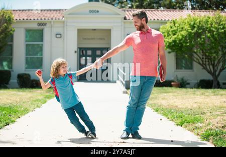 Il genitore e l'allievo divertente della scuola primaria vanno di pari passo. Insegnante in t-shirt e carino scolaro con zaino vicino al parco scolastico. Foto Stock