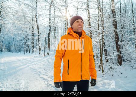 Corridore sorridente di mezza età vestito da giacca antivento arancione brillante, che corre lungo la pittoresca foresta innevata. Persone sportive attive e vincenti Foto Stock