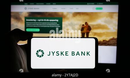 Persona che detiene un telefono cellulare con il logo della società finanziaria danese Jyske Bank sullo schermo di fronte alla pagina Web aziendale. Mettere a fuoco il display del telefono. Foto Stock