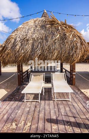 Due sedie a sdraio vuote sotto il tetto di paglia su un ponte di legno sulla spiaggia sabbiosa, con luci a corde che pendono sopra il cielo blu di Curacao Foto Stock