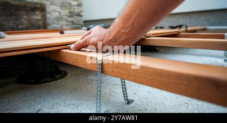 Vista ad angolo basso della mano maschile che posiziona un distanziatore di plastica tra le tavole di legno mentre sta costruendo un patio esterno rinnovato su un edificio di terreno Foto Stock