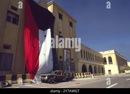 Prima guerra del Golfo: 8 marzo 1991 Un'enorme bandiera del Kuwait, forata da fori di conchiglia, copre il muro di un edificio a Seif Palace, casa dell'Emiro a Kuwait City. Accanto alla bandiera c'è un grande ritratto piastrellato di Saddam Hussein che indossa un costume da lavoro, deturpato da proiettili. Foto Stock