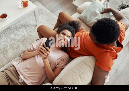 Vista dall'alto di una giovane donna africana che usa lo smartphone e sorride mentre passa del tempo a casa con il suo ragazzo Foto Stock