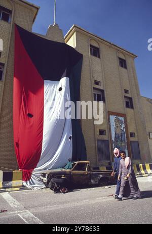 Prima guerra del Golfo: 8 marzo 1991 due kuwaitiani camminano davanti a un'enorme bandiera del Kuwait, forata da fori di conchiglia, e un ritratto sfacciato di Saddam Hussein sul muro di un edificio, parte del Seif Palace, casa dell'emiro a Kuwait City. Foto Stock