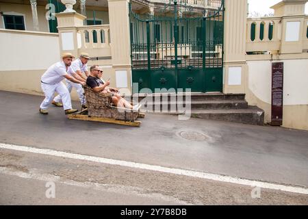 Turisti e turisti che si divertono facendo un giro sulle tradizionali slitte da slittino con cesto di vimini a Monte, Funchal, sull'isola portoghese di Madeira Foto Stock