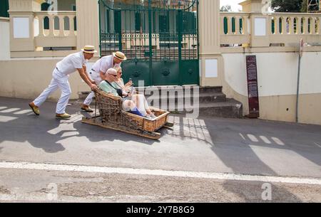 Turisti e turisti che si divertono facendo un giro sulle tradizionali slitte da slittino con cesto di vimini a Monte, Funchal, sull'isola portoghese di Madeira Foto Stock