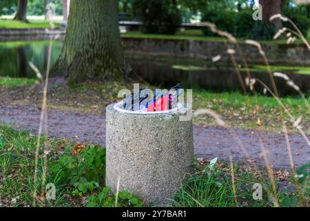 Un bidone di rifiuti in cemento pieno di sacchetti blu e neri per rifiuti canini si trova accanto al sentiero davanti al fossato pieno d'acqua del castello di Mellenthin. Schlossallee, Usedom-Süd, Meclemburgo-Pomerania Occidentale, Germania. Foto Stock