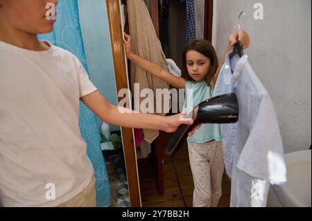 Una giovane ragazza aiuta il fratello ad asciugare i vestiti con un asciugacapelli. Vita domestica e lavoro di squadra durante le faccende domestiche. Foto Stock