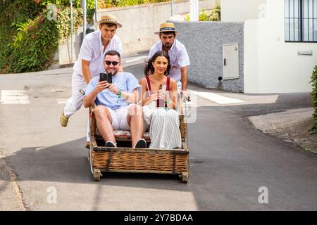 Turisti e turisti che si divertono facendo un giro sulle tradizionali slitte da slittino con cesto di vimini a Monte, Funchal, sull'isola portoghese di Madeira Foto Stock