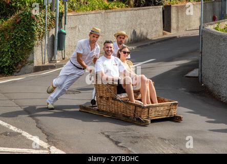 Turisti e turisti che si divertono facendo un giro sulle tradizionali slitte da slittino con cesto di vimini a Monte, Funchal, sull'isola portoghese di Madeira Foto Stock