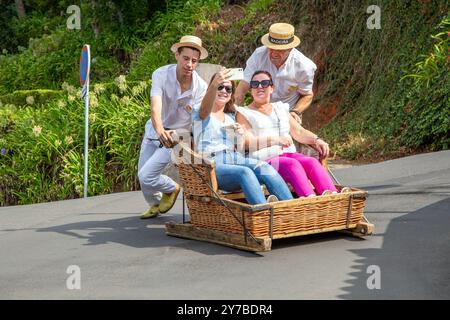 Turisti e turisti che si divertono facendo un giro sulle tradizionali slitte da slittino con cesto di vimini a Monte, Funchal, sull'isola portoghese di Madeira Foto Stock