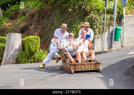 Turisti e turisti che si divertono facendo un giro sulle tradizionali slitte da slittino con cesto di vimini a Monte, Funchal, sull'isola portoghese di Madeira Foto Stock