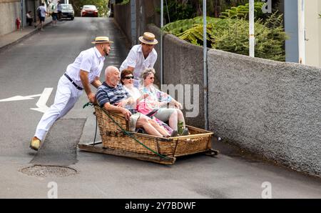 Turisti e turisti che si divertono facendo un giro sulle tradizionali slitte da slittino con cesto di vimini a Monte, Funchal, sull'isola portoghese di Madeira Foto Stock