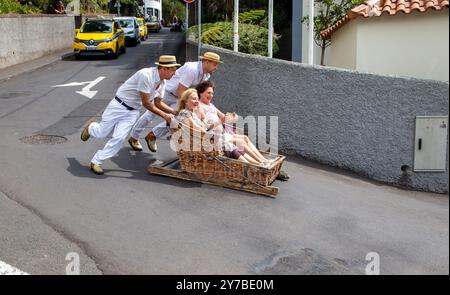 Turisti e turisti che si divertono facendo un giro sulle tradizionali slitte da slittino con cesto di vimini a Monte, Funchal, sull'isola portoghese di Madeira Foto Stock