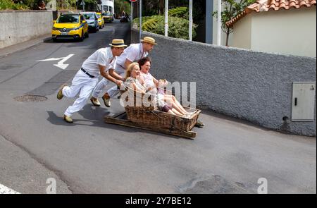 Turisti e turisti che si divertono facendo un giro sulle tradizionali slitte da slittino con cesto di vimini a Monte, Funchal, sull'isola portoghese di Madeira Foto Stock