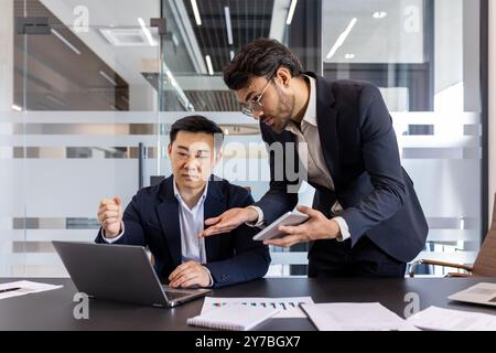 Due professionisti impegnati in discussioni strategiche in uffici moderni. Uno seduto utilizzando un computer portatile e un altro che mostra le informazioni sul telefono. Concetto di lavoro di squadra, collaborazione, tecnologia nel business Foto Stock