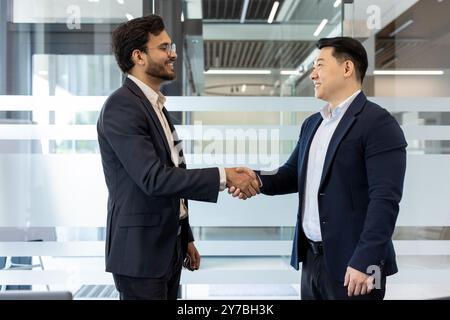 Due uomini d'affari si scambiano una stretta di mano sicura in un ambiente d'ufficio moderno, mostrando interazione professionale e lavoro di squadra. Si alzano l'uno di fronte all'altro, indossando un abbigliamento formale Foto Stock