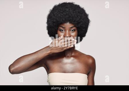 Ritratto di una bella giovane donna africana che copre la bocca con la mano mentre si trova su sfondo grigio Foto Stock