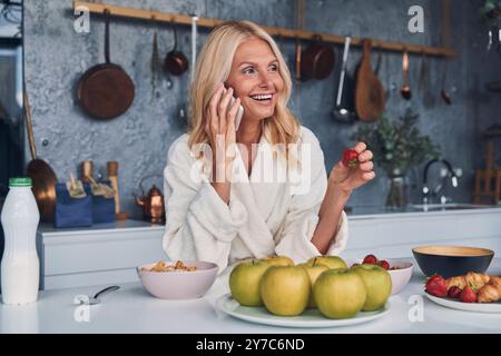 Bella donna matura in accappatoi che parla al telefono cellulare e sorride mentre fa colazione nella cucina domestica Foto Stock