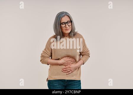 Donna matura frustrata che soffre di mal di stomaco mentre si trova su sfondo grigio Foto Stock
