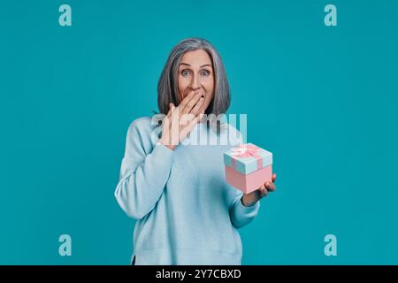Bella donna matura che tiene in mano la confezione regalo e copre la bocca mentre si sta in piedi su sfondo blu Foto Stock