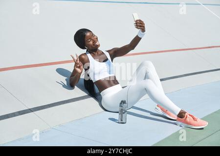 Bella giovane donna africana in abbigliamento sportivo che fa videochiamate e sorride mentre siede su una pista sportiva all'aperto Foto Stock