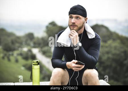 Atleta muscoloso con barba con asciugamano che controlla le calorie bruciate sullo smartphone dopo una buona sessione di allenamento nel parco cittadino Foto Stock