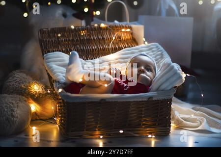 Un bambino appena nato con abiti natalizi che dorme in un cestino sul pavimento con decorazioni natalizie. Capodanno Foto Stock