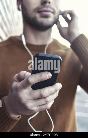 Uomo sorridente ascoltando musica su cuffie e appoggiata contro un muro di mattoni. Ritratto di uomo sorridente con le cuffie e il cellulare in piedi dal mattone w Foto Stock