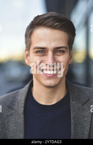 Un uomo alla moda e felice soggiorna in strada con un cappotto grigio e un maglione blu Foto Stock