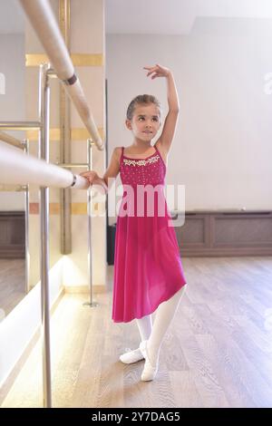 L'affascinante bambina sogna di diventare una ballerina. La ragazza con il vestito rosa sta ballando, tenendo il bar. La bambina sta studiando il balletto Foto Stock