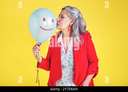 Donna anziana, palloncino e studio per festeggiare, emoji per il viso felice e baci su sfondo giallo. Persona femminile, festa di pensionamento ed evento per Foto Stock