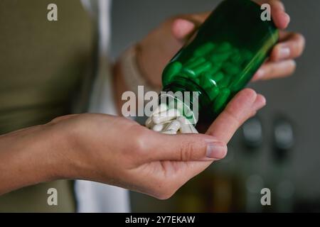 Primo piano di una donna che versa capsule bianche dal biberon verde alla mano. La persona sta assumendo pillole o vitamine. Concetto di integratori, medicina o salute c Foto Stock