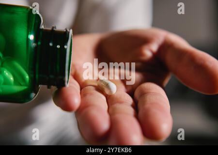 Primo piano di un uomo che versa capsule bianche dal flacone verde alla mano. La persona sta assumendo pillole o vitamine. Concetto di integratori, medicina o auto di salute Foto Stock