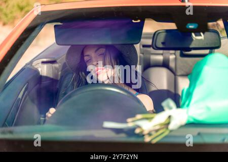 Una donna sta guidando un'auto con un mazzo di fiori sul sedile posteriore. Indossa un cappello e ha gli occhi chiusi. Foto Stock