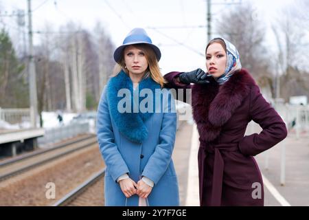 Due donne che indossano cappotti e cappelli in pelliccia si trovano su una piattaforma ferroviaria. Uno di loro ha in mano una borsetta Foto Stock