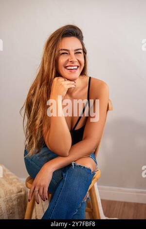 Una giovane donna con i capelli lunghi sorridente mentre siede su uno sgabello di legno, indossa un top nero e jeans strappati, all'interno. Foto Stock