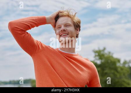 Giovane uomo rosso felice che tiene per mano i capelli e sorride mentre si trova all'aperto Foto Stock