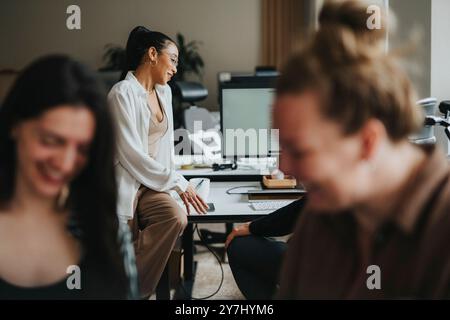 Giovane donna d'affari che si appoggia alla scrivania e parla con un collega sul posto di lavoro Foto Stock