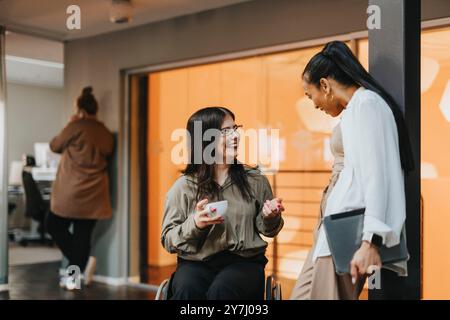 Imprenditrice felice con disabilità fisica che tiene una tazza di caffè e discute con la collega in carica Foto Stock