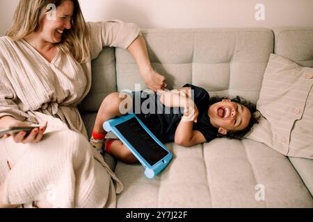 Mamma che solletica il figlio sdraiato sul divano con un tablet digitale a casa Foto Stock