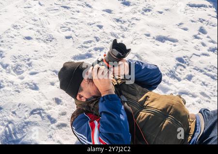 un uomo anziano con una macchina fotografica in mano, sdraiato sulla neve e fotografato un paesaggio invernale di montagna. Foto Stock