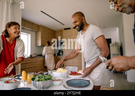 I membri della famiglia felici si aiutano a vicenda per preparare un tavolo da pranzo a casa Foto Stock