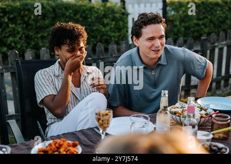 Giovane che copre la bocca con le mani mentre si siede accanto a un amico maschio a cena nel cortile posteriore Foto Stock